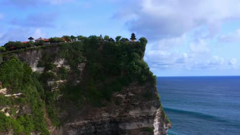 Approaching-aerial-view-of-Uluwatu-temple-on-a-tall-cliff-near-the-ocean-in-Bali,-Indonesia