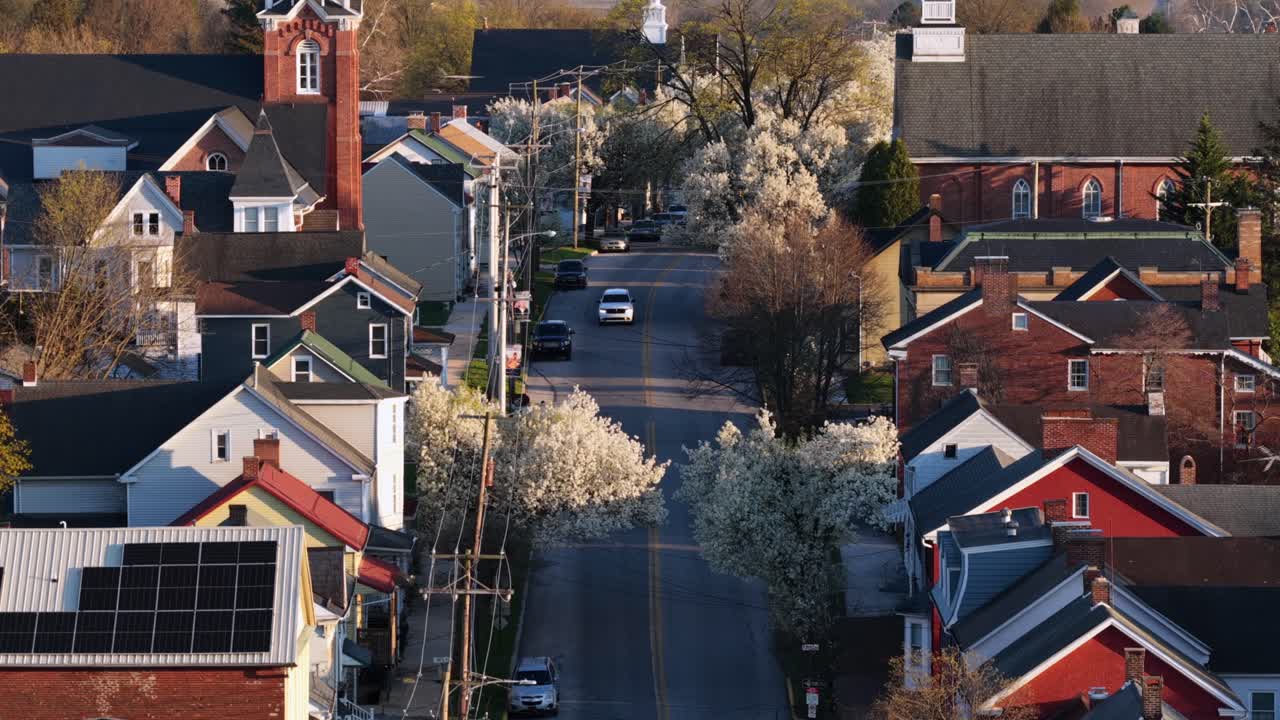 Zoom aereo su un tranquillo quartiere della Pennsylvania all'ora d'oro, che mostra tipiche case a schiera americane, alberi primaverili in fiore, pannelli solari sui tetti e una tranquilla strada residenziale.