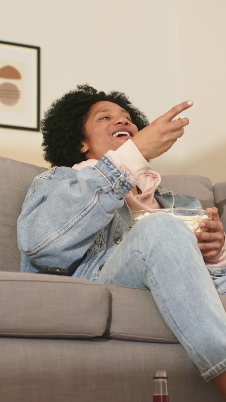 Video verticale: Uomo in giacca di jeans e felpa con cappuccio che preme il telecomando e reagisce alla TV a casa con popcorn.