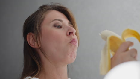 close up of lady carefully eating banana under bright bulb light creating soft illumination highlighting texture of fruit and her gentle facial expression in clean bright indoor environment