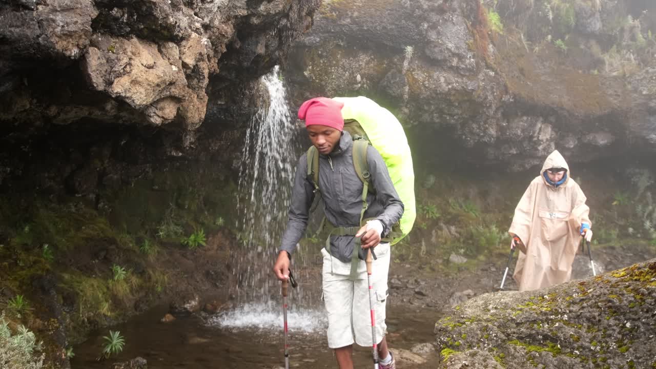los excursionistas caminan con mochilas y palos de senderismo mientras caminan por el monte kilimanjaro más allá de las cataratas