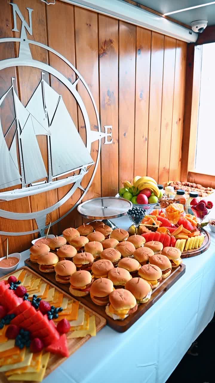 Tavolo da buffet catering con stuzzichini e decorazioni murali a tema nave. Antipasti assortiti inclusi mini-burger, piatto di salumi e torta su tovaglia bianca.