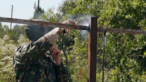 artesano con máscara suelda el riel metálico de la valla en un día de verano