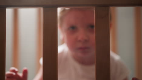 upset blond toddler looks out of crib grabbing wooden planks