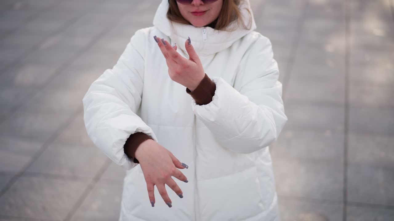 Down up view of student in white winter coat and head warmer wearing black shield glasses making excited hand dance moves on tiled outdoor pavement under pale winter sky with snow backdrop