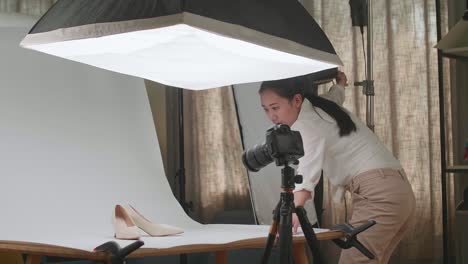 asian female photographer adjusting light equipment to get good illumination on women's shoes while taking photos of the products in home studio