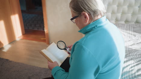 senior woman in teal sweater sitting on bed, holding a magnifying glass to read a hardcover book. sunlight fills the room, creating a peaceful and focused atmosphere