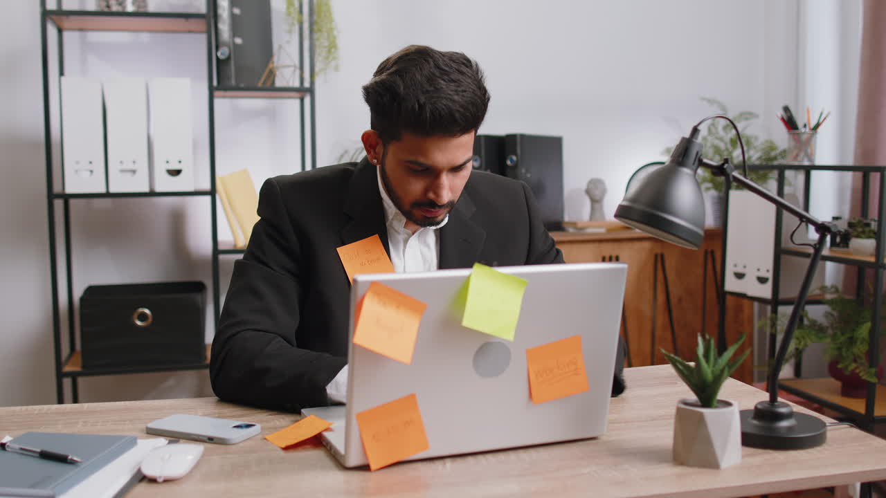 Tired sad exhausted businessman working on laptop at office with many sticker tasks panic attack