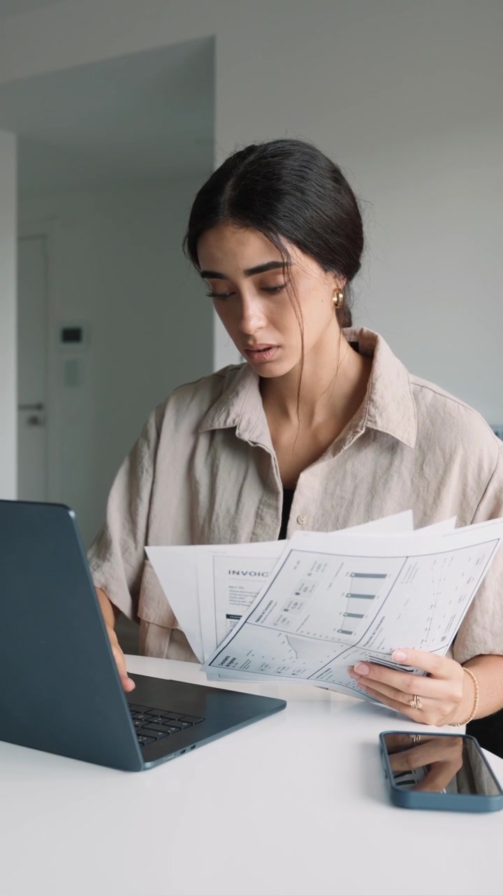Giovane donna che esamina documenti finanziari e lavora al laptop.