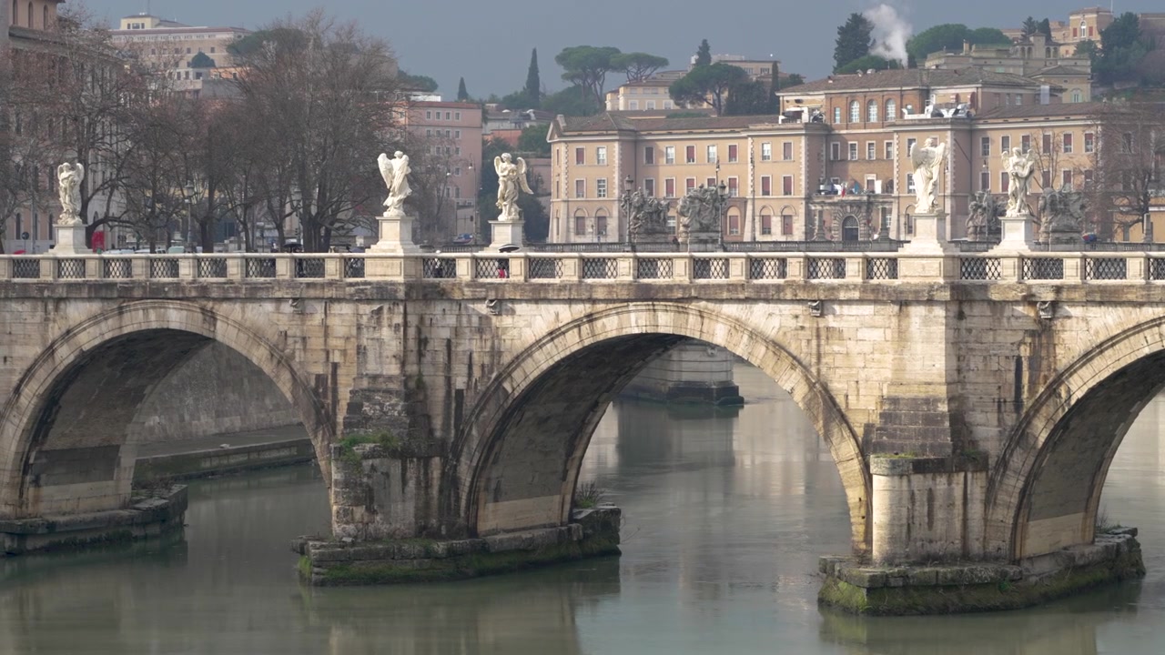 Statue di angeli fiancheggiano Ponte Sant'Angelo mentre i suoi archi si estendono su acque ancora verdi con isolati cittadini dai colori tenui nella morbida luce invernale