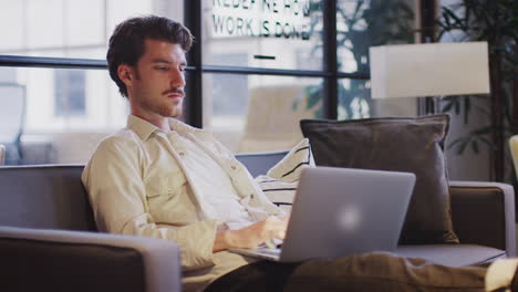 Un-Joven-Trabajando-En-Una-Computadora-Portátil-Sentado-Con-Los-Pies-En-Alto-En-Una-Sala-De-Estar-De-Oficina,-Vista-Lateral