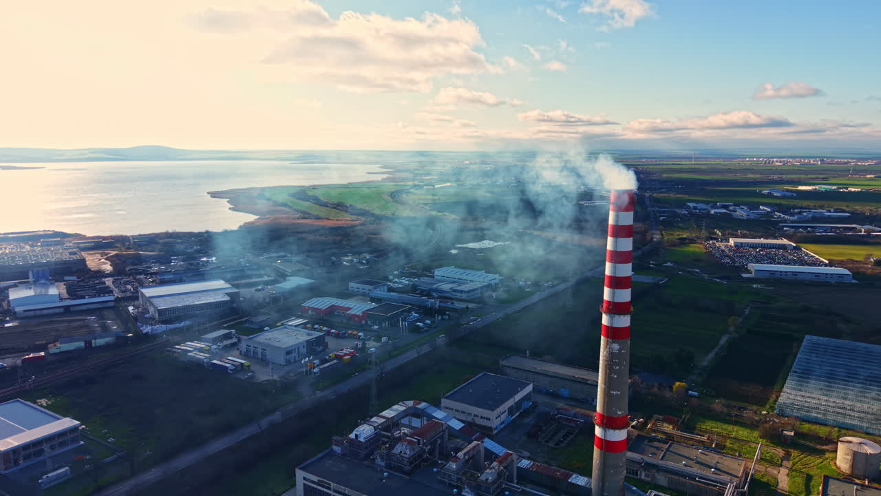 Il fumo sale dalla ciminiera di una fabbrica vicino a uno specchio d'acqua, mentre il sole splende sulla zona industriale al mattino presto
