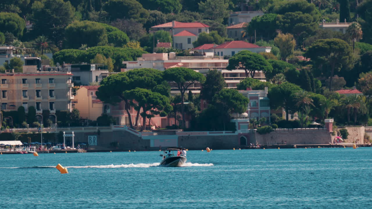 A sleek motorboat speeds across the clear blue water with coastal buildings and lush trees in the background