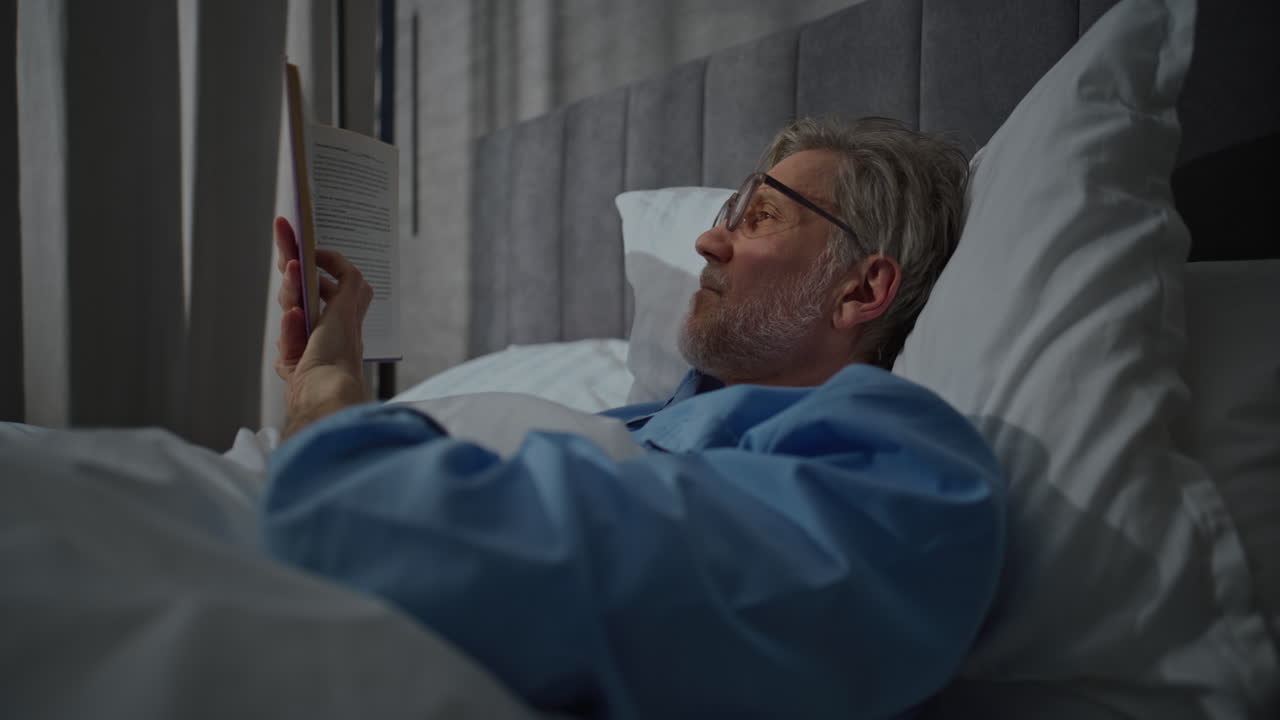Waking Up in Morning in Bright Cozy Bedroom Mature Man Lies Relaxed in White Bed Reading Book