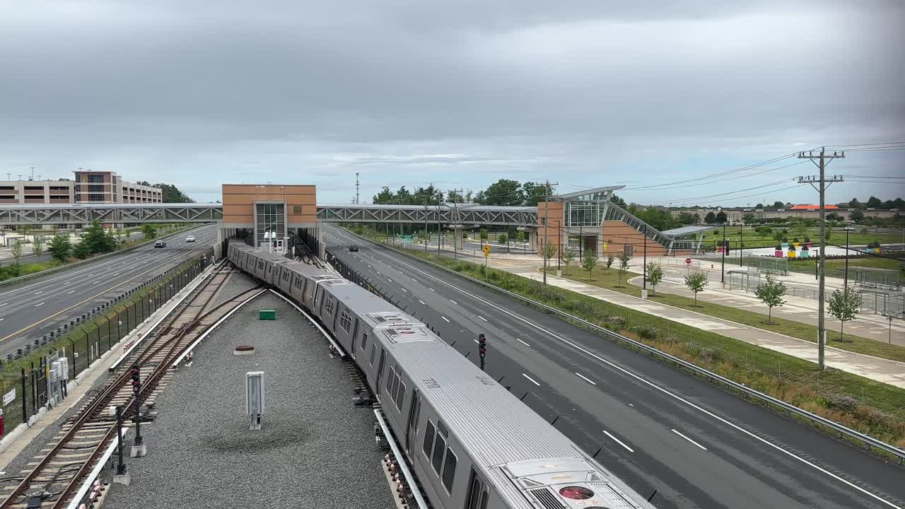 Train leaving modern metro station located between lanes of divided highway with skybridge, sidewalks, traffic lanes, trees, streetlights, and overcast sky in suburban West Virginia environment