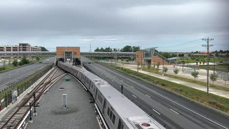 train leaving modern metro station located between lanes of divided highway with skybridge, sidewalks, traffic lanes, trees, streetlights, and overcast sky in suburban west virginia environment