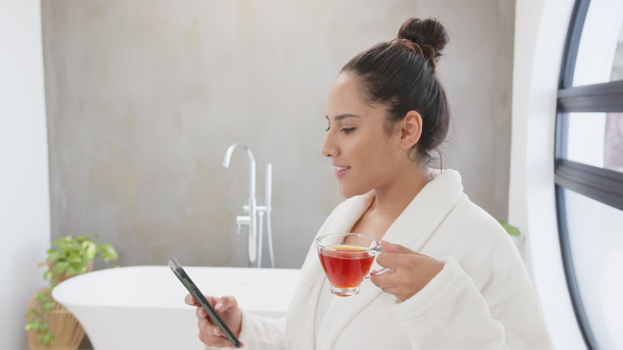Donna asiatica che tiene lo smartphone e beve tè, rilassandosi in un bagno spa a casa.