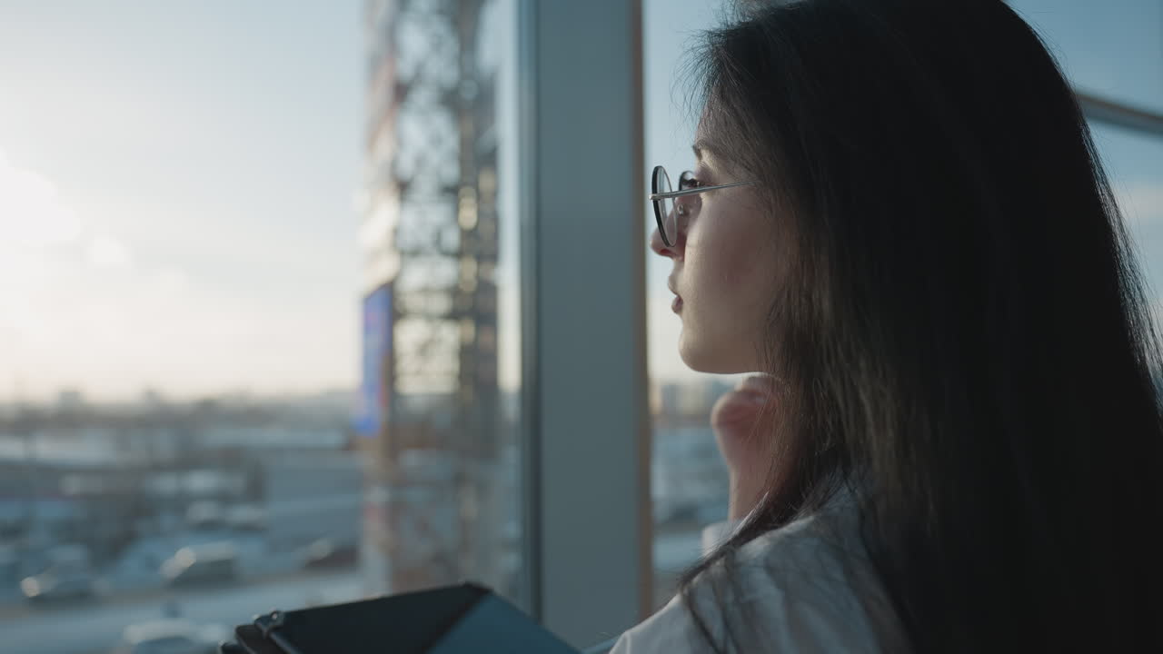 Close up of professional woman adjusting glasses with one hand while holding tablet in other, facing sunlit city view through window, natural light softly illuminating face and background