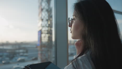 close up of professional woman adjusting glasses with one hand while holding tablet in other, facing sunlit city view through window, natural light softly illuminating face and background