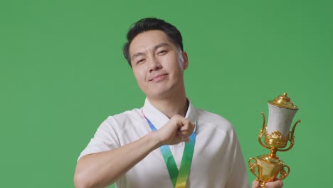 close up of asian man with a gold medal showing gold trophy to camera, smiling, and pointing himself being proud winning as the first winner on green screen background in the studio