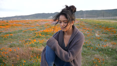 una chica joven y bonita sentada en una roca en un campo de flores silvestres que se ve triste y sola con el pelo negro soplando en el viento en cámara lenta