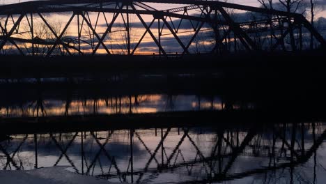 a little red, arched, steel truss bridge over a river at dusk with reflections in the water and cars driving by in small town america