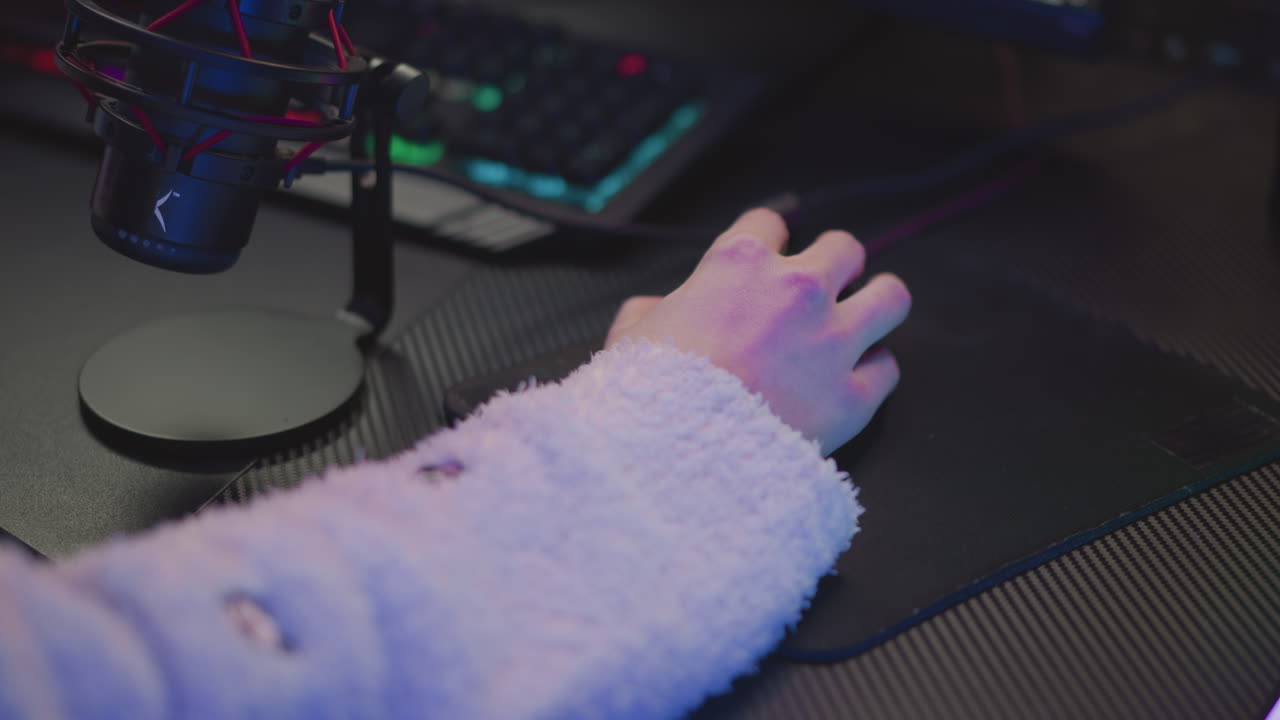 Close up hand of person in fluffy sweater using mouse on dark textured desk with RGB keyboard and microphone visible, subtle pink and blue ambient lighting sets cozy digital workspace