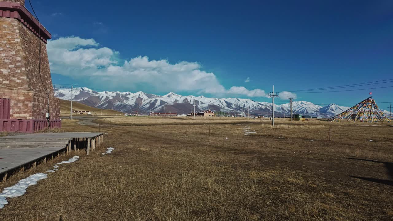 Qilian Grassland, Xining, Qinghai Province, China - A Wide Grassland Village Sits Beneath Snow-capped Mountains, with Colorful Prayer Flags Standing Out Against the Clear Blue Sky - Pan Right Shot