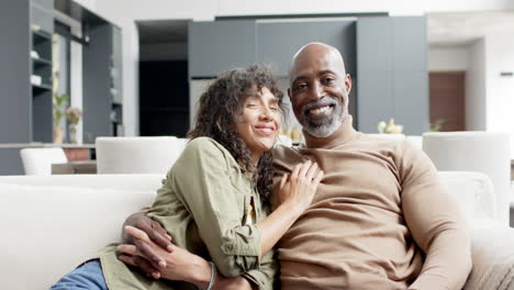 Portrait-of-happy-biracial-couple-embracing-and-sitting-on-couch-at-home,-slow-motion