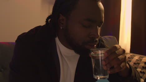 man sitting on sofa after drinking alcohol at house party drinking glass of water for hangover