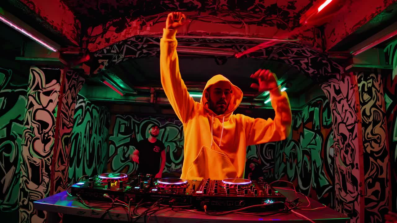 Neon ışıklı, grafitilerle kaplı bir yeraltı kulübünde DJ'in geniş açılı çekimi, canlı, kentsel bir atmosfer oluşturan.