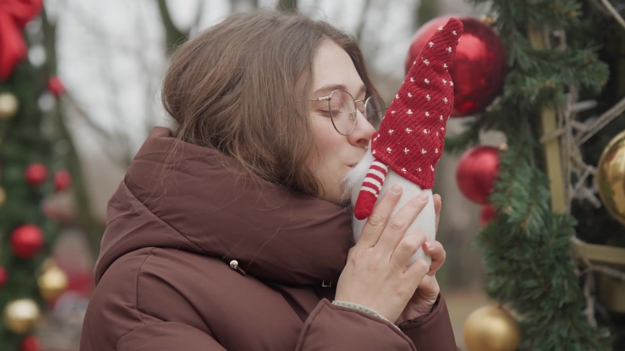 Jovem mulher examina gnomo vermelho, sorrindo com casaco puffer marrom perto de um arco de Natal decorado com bolas douradas e vermelhas, guirlanda de folhagem e neve suave, momento espontâneo de mercado transmitindo um feriado aconchegante