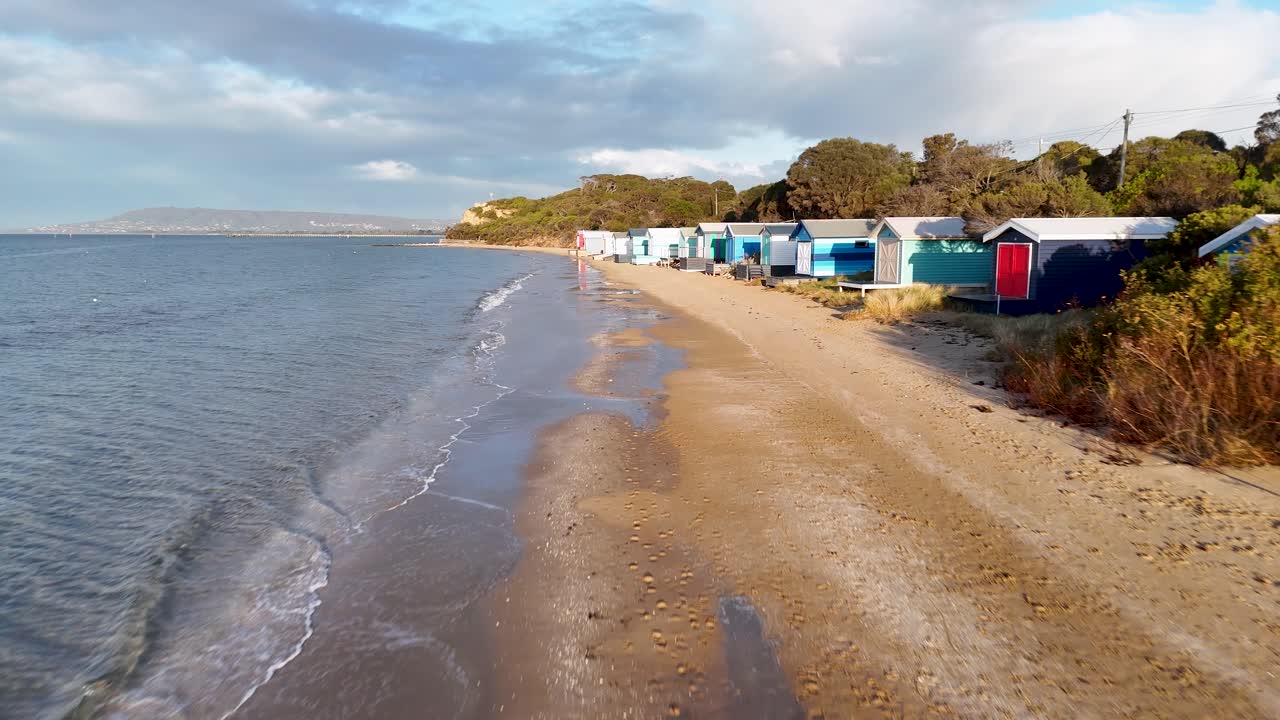 Drone glides above sandy beach, passing vibrant bathing boxes under soft morning sunlight, calm atmosphere