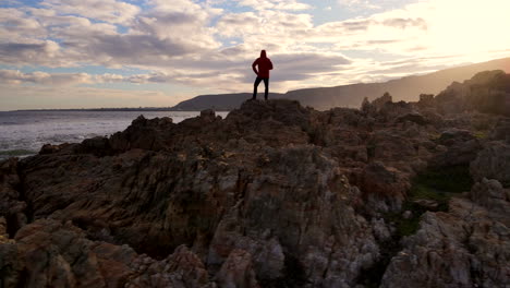 hombre en la costa rocosa mirando hacia el océano, el sol se pone héroe aéreo disparado