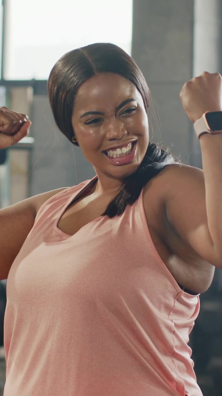 Vertical video: Cheering woman in gym after set, flexing arms and showing pink tank and smartwatch
