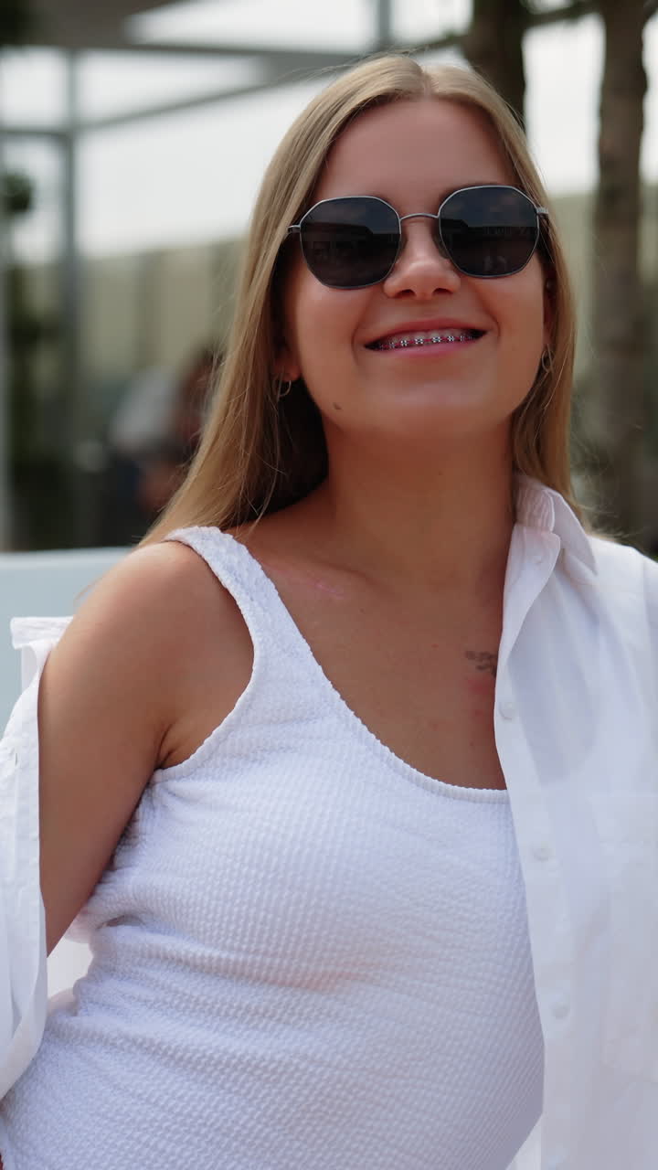 Blonde girl sitting on deckchair in white clothes wearing sunglasses. Smiling woman covers her shoulder from sun. Vertical video
