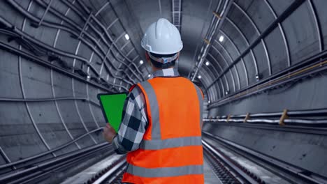 back view of asian male engineer with safety helmet working on a green screen tablet and looking around while standing in underground subway tunnel