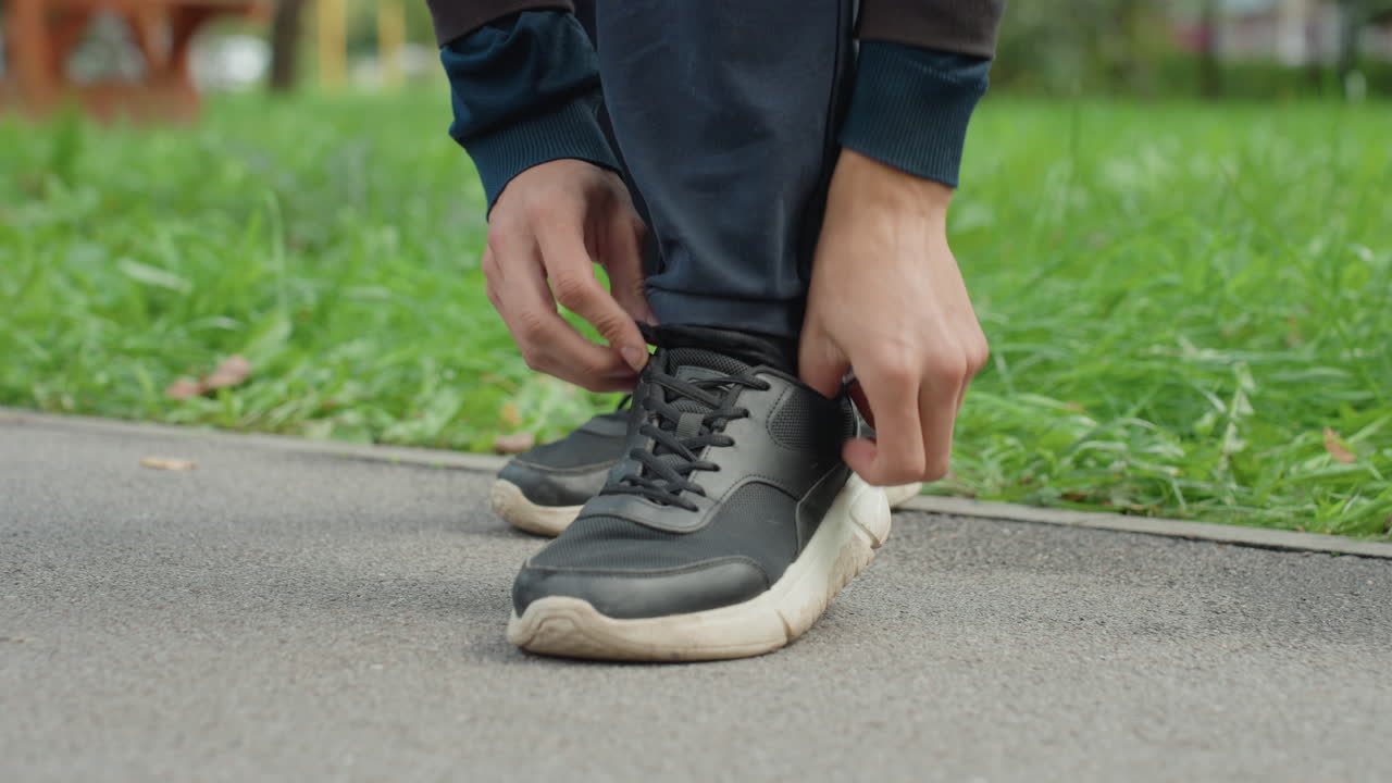 uomo che si allaccia le scarpe da ginnastica nel parco, dettaglio ravvicinato da una bassa angolazione delle mani che sistemano scarpe da corsa nere sull'asfalto accanto all'erba verde, jeans casual, suola testurizzata, tenue luce mattutina, atmosfera cinematografica broll