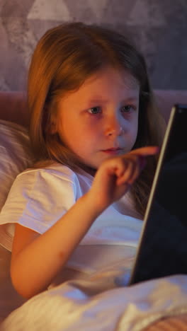 little girl uses large tablet computer touching screen to play development game on bed with pillows at night lamp light in shaded room in evening slow motion