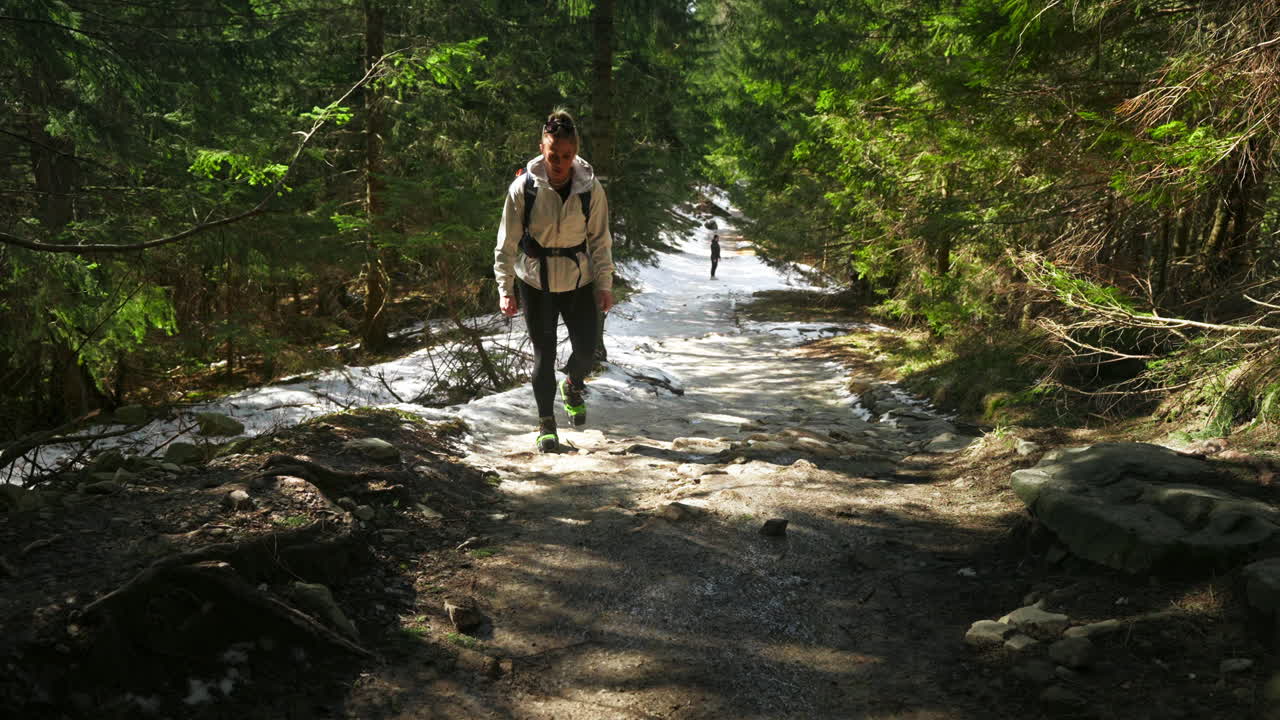 A woman walks on a narrow forest trail surrounded by trees and dappled mountain light, slowly hiking on muddy trail in shade