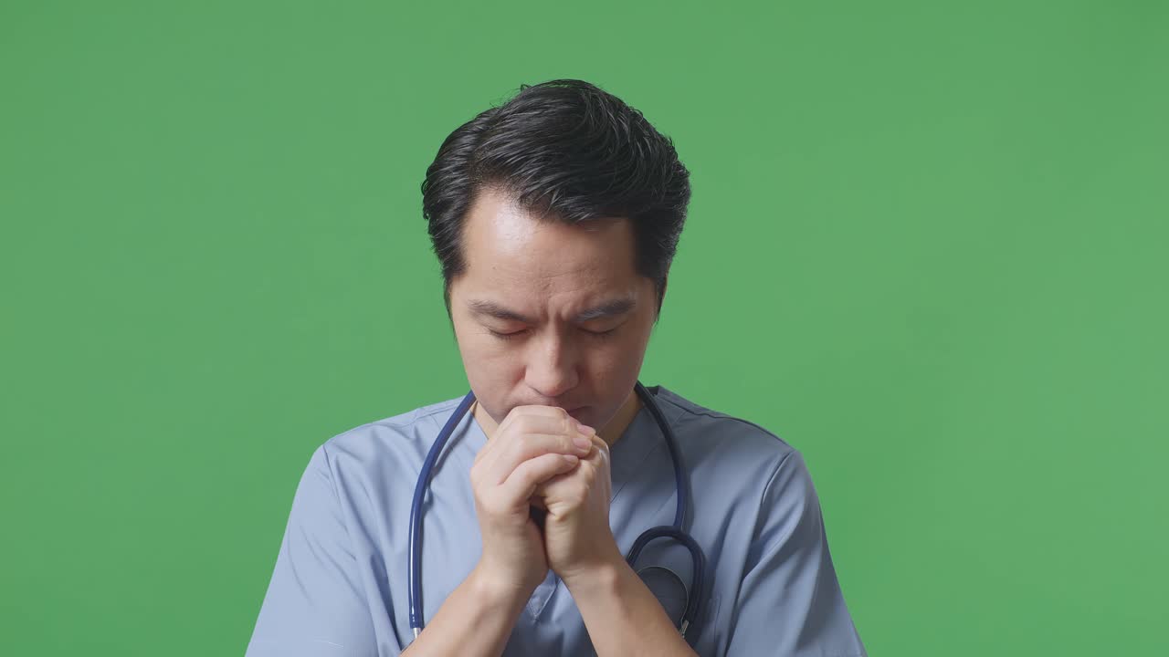스테토스코프를 들고 있는 아시아 남성 의사의 클로즈업은 병원의 녹색 스크린 배경에서 무언가를 위해 기도합니다.