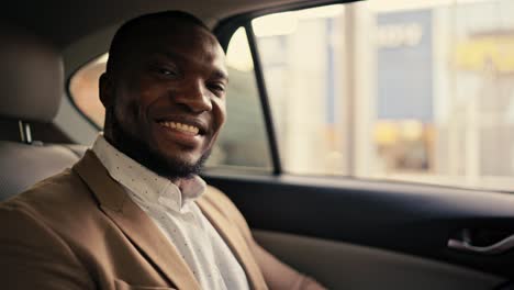 Portrait-of-a-happy-man-with-Black-skin-with-a-beard-in-a-brown-jacket-who-sits-in-the-back-seat-of-a-modern-car-smiling-and-looking-at-the-camera-during-his-business-trip-in-the-city