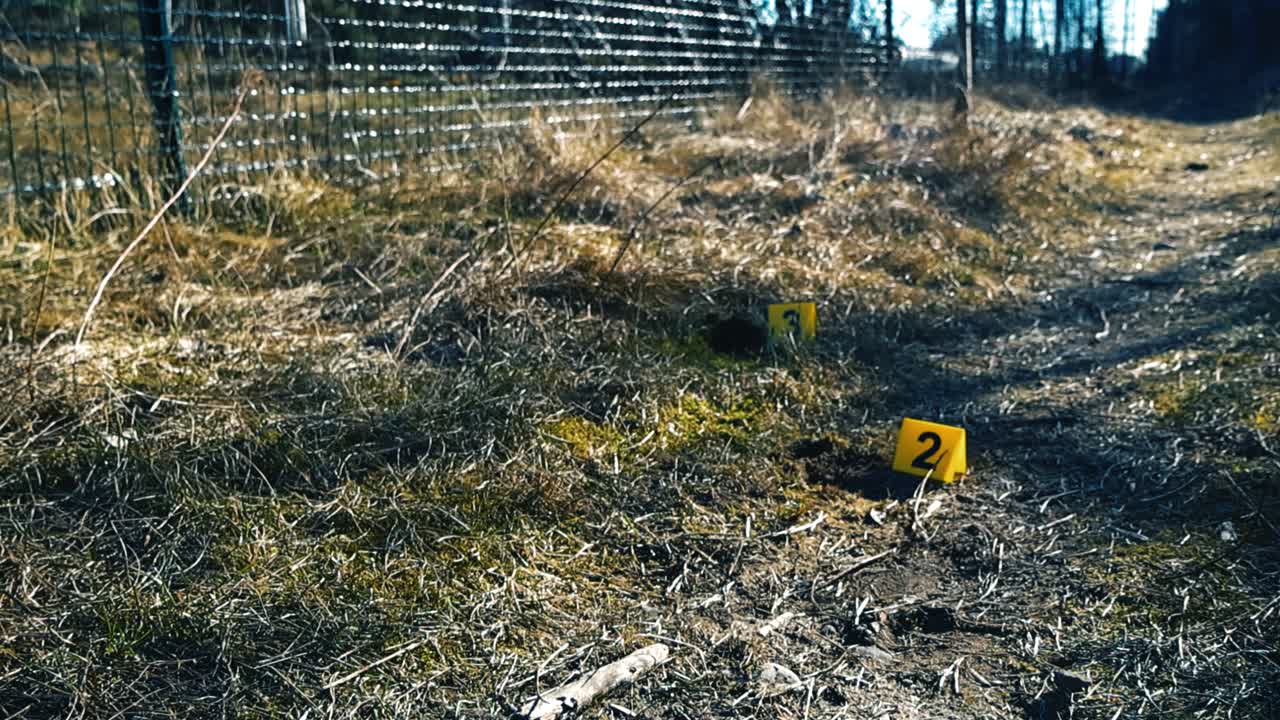 Yellow forensic markers with numbers indicating evidence at an outdoor crime scene next to a dirt road and a fence, symbolizing a police investigation into a serious criminal act