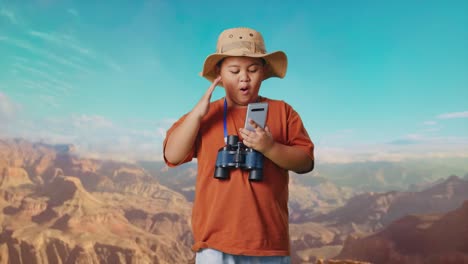 asian boy with a hat and binoculars looking at smartphone then saying wow. boy researcher examines something while traveling at the top of mountain, travel tourism adventure concept