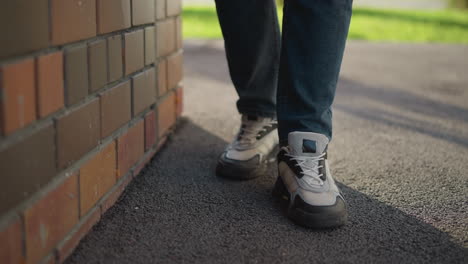 close up leg view of person in sneakers and denim jeans standing beside brick wall with shadow stretching on pavement, scene features casual posture with soft sunlight and green background