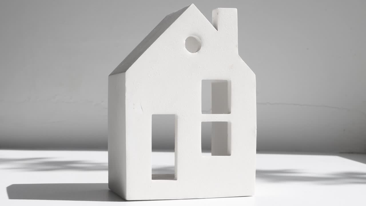 Close-up of a ceramic figurine in the form of a house with windows and a doorway on a light gray background with a shadow from a plant.