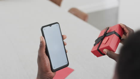 african american man holding a smartphone and a gift box at home with copy space, on a video call