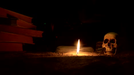 wizard's desk. a desk lit by candle light. a human skull, old books on sand surface. halloween still-life background with a different elements on dark toned foggy background. slider shot