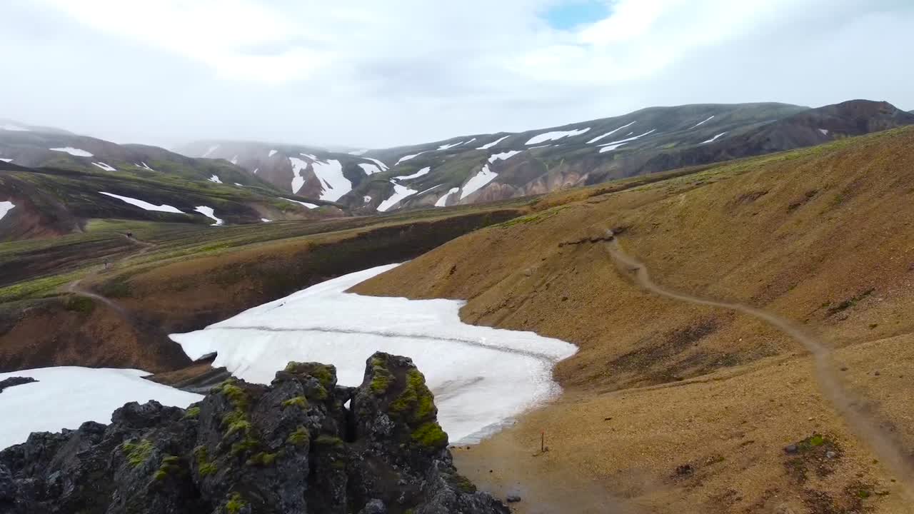 Aerial drone fotoage flying over and gliding towards large vast snow covered Iceland volcanic terrain mountains during a cloudy day. Sharp volcanic rocks covered in moss in foreground, hiking trail.