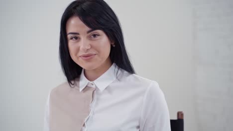 portrait of a beautiful dark-haired girl in studio. young woman in white blouse and short skirt sitting on the chair with laptop on light background.